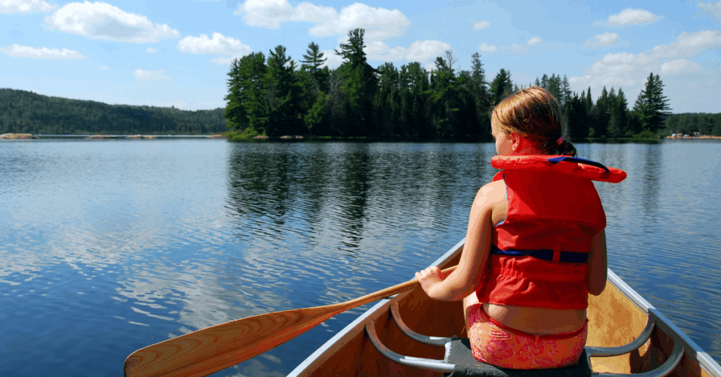 A young girl paddling a canoe alone on a calm lake, viewed from behind